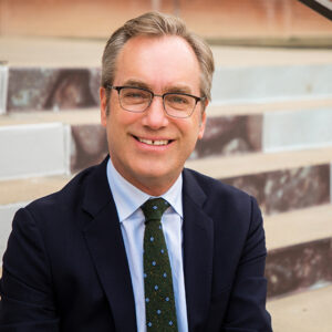 A man with short blond hair with glasses wearing a blue button down shirt, dark green tie and navy blazer sitting on steps.